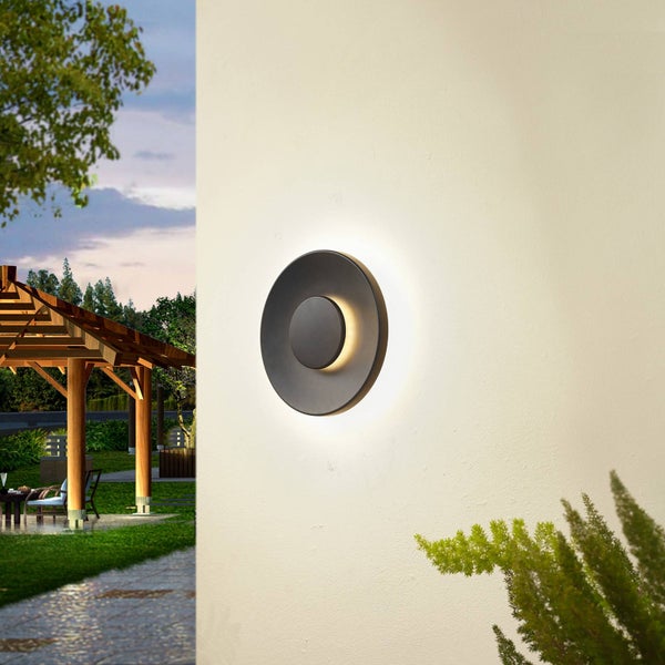 Runde schwarze LED-Außenwandleuchte an einer hellen Wand, im Hintergrund ein Garten mit Holzpavillon und Sitzbereich bei Dämmerung.