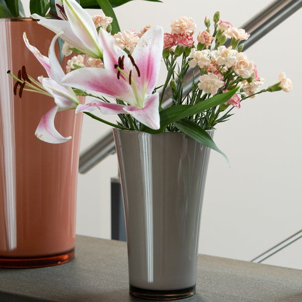 Graue glänzende Blumenvase in konischer Form mit rosa Lilien und Nelken auf einer grauen Fläche in einem hellen Raum.