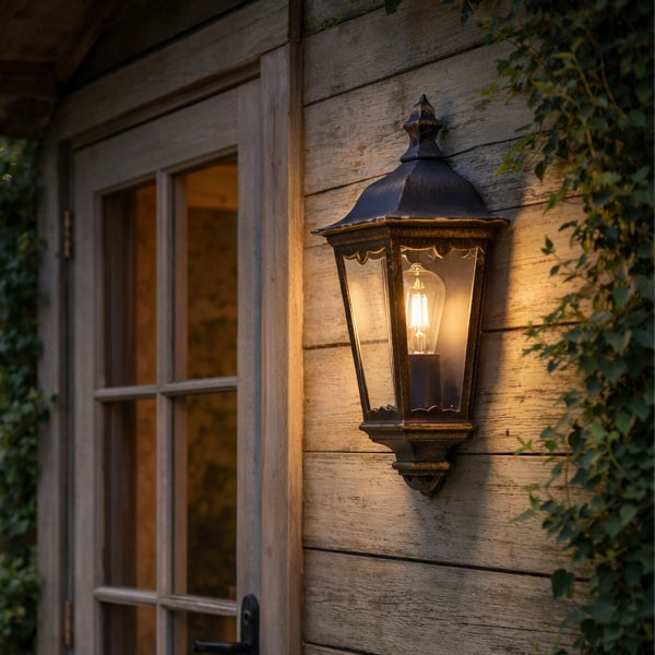 Klassische Außenwandleuchte in Laternenform aus Metall und Glas, montiert an einer Holzwand neben einer Tür, beleuchtet mit warmem Licht.