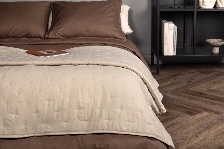 Beige gesteppte Tagesdecke auf einem Bett mit braunen Kissen neben einem schwarzen Regal auf Fischgrätparkett.