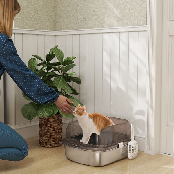 Moderne Katzentoilette mit transparenter Haube und Streuschaufel, eine Katze steht darin neben einer knienden Frau in einem hellen Raum.