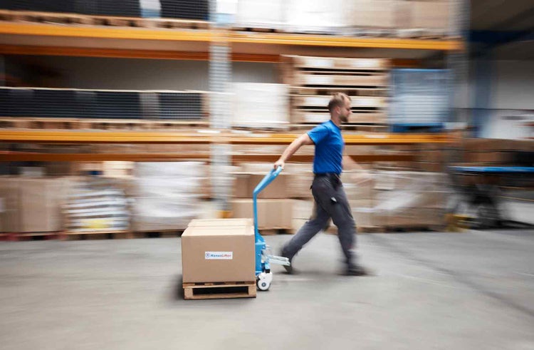 Ein Mitarbeiter zieht einen Hubwagen mit einem Karton auf einer Palette durch ein Lager mit hohen Regalen.