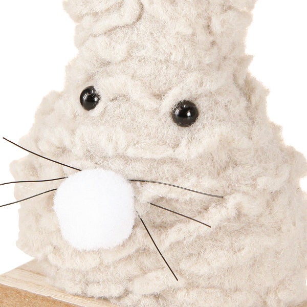 Osterhasenfigur aus flauschigem Textilmaterial mit schwarzen Knopfaugen und Schnurrhaaren auf einem Holzsockel