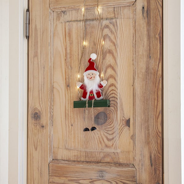 Dekorativer Weihnachtsmann auf einer Schaukel mit Lichtern an einer Holztür.