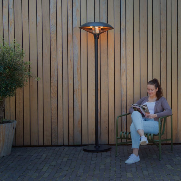 Schwarzer Infrarot-Terrassenheizer in Betrieb auf einer Terrasse neben einer sitzenden Person vor einer vertikalen Holzwand.
