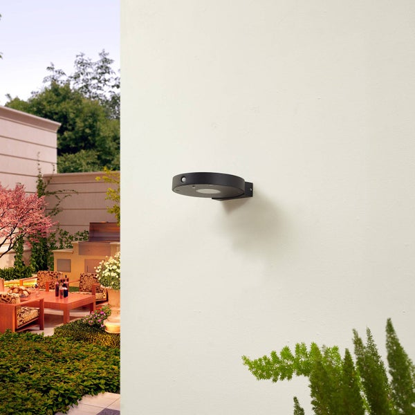 Schwarze runde Außenwandleuchte mit Bewegungsmelder an einer Wand in einem hellen Garten mit Terrasse, Holzmöbeln und Pflanzen.