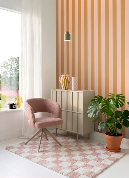 Hell gestalteter Wohnraum mit gestreifter Tapete, beigefarbenem Schrank, rosa Sessel, kariertem Teppich und einer Monstera-Pflanze.