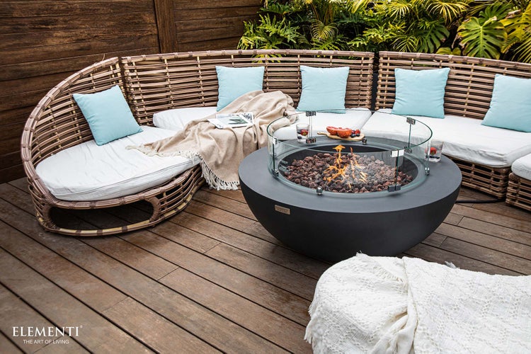 Moderner Loungebereich im Freien mit einem runden Gasfeuertisch in Betonoptik, einem geschwungenen Rattansofa und einer Holzterrasse.