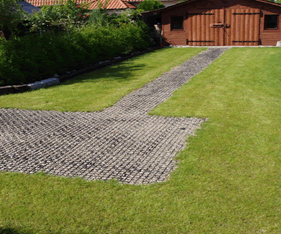 Rasengitter aus Kunststoff mit Kiesfüllung als Einfahrt und Gartenweg auf einer Rasenfläche vor einem Holzhaus.