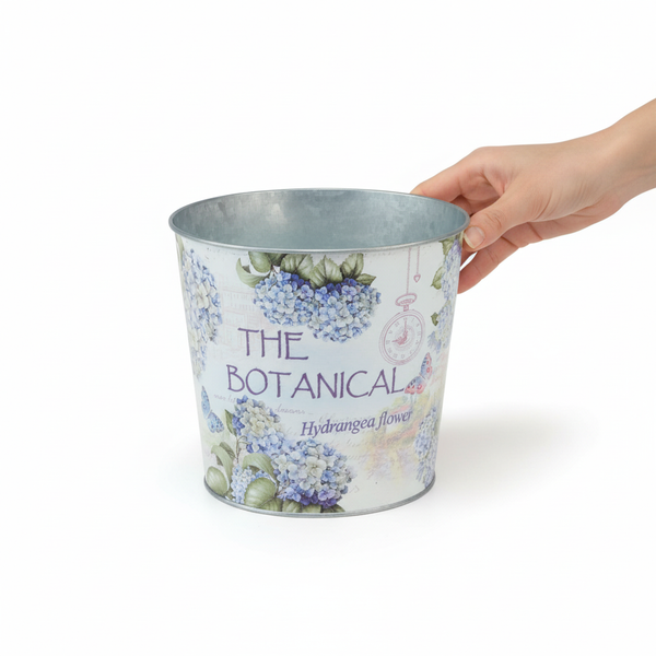 Dekorativer Übertopf aus Metall mit Hortensien-Motiv und Schriftzug The Botanical. Eine Hand hält den Rand zur Größenanzeige.