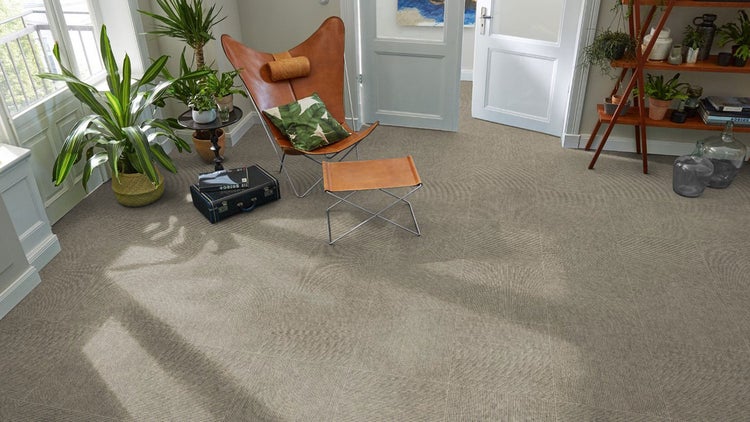 Heller Wohnraum mit strukturierten Teppichfliesen, einem Butterfly-Sessel aus Leder, grünen Zimmerpflanzen und einer weißen Tür.