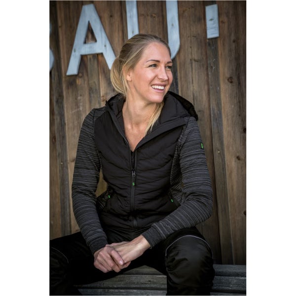 Frau in schwarz-grauer Hybrid-Arbeitsjacke mit Kapuze und schwarzer Arbeitshose vor einer Holzwand.
