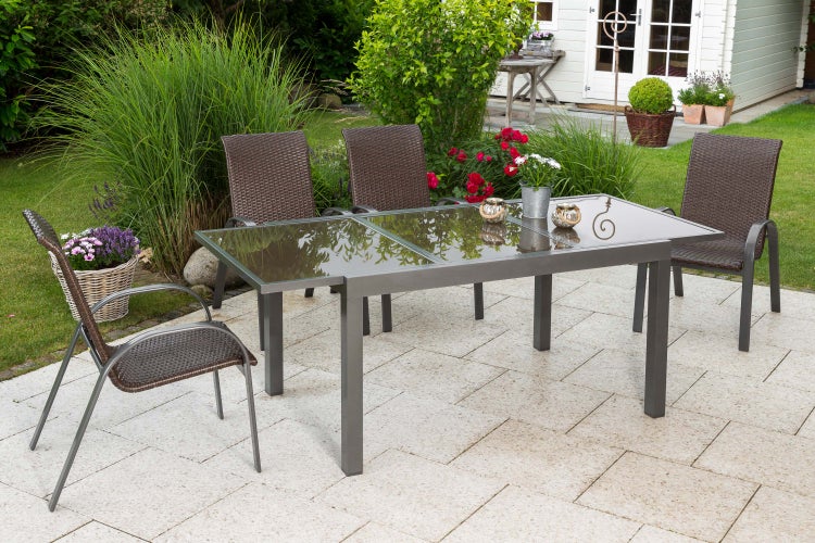 Gartenmöbel-Set mit rechteckigem Glastisch und vier dunklen Geflechtsesseln auf einer hellen Steinterrasse im Garten.