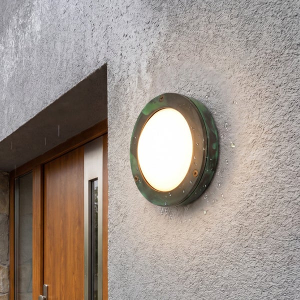 Runde Außenwandleuchte in patinierter Optik an einer Hauswand neben einer Holztür.