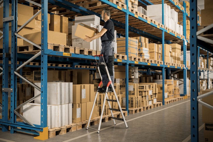 Mann auf einer Trittleiter in einem Lager, der eine Kiste trägt