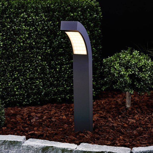 Moderne LED-Wegeleuchte in Anthrazit, beleuchtet in einem Gartenbeet mit Rindenmulch und grünen Hecken.