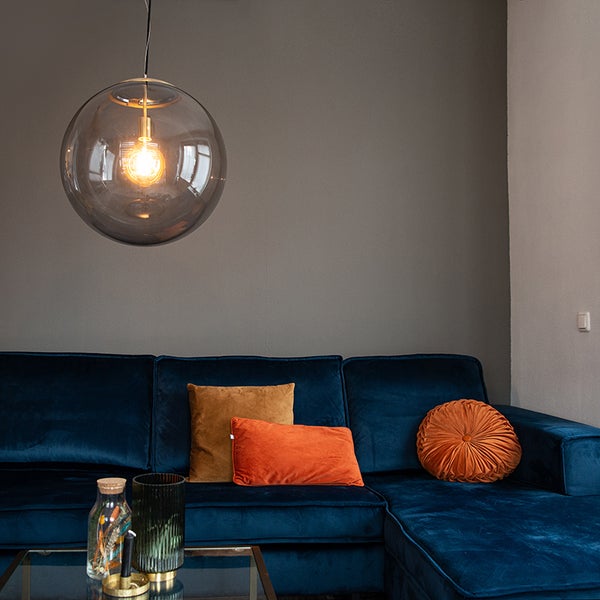 Modernes Wohnzimmer mit großer Pendelleuchte aus Rauchglas über einem dunkelblauen Samtsofa mit orangefarbenen und senfgelben Kissen.