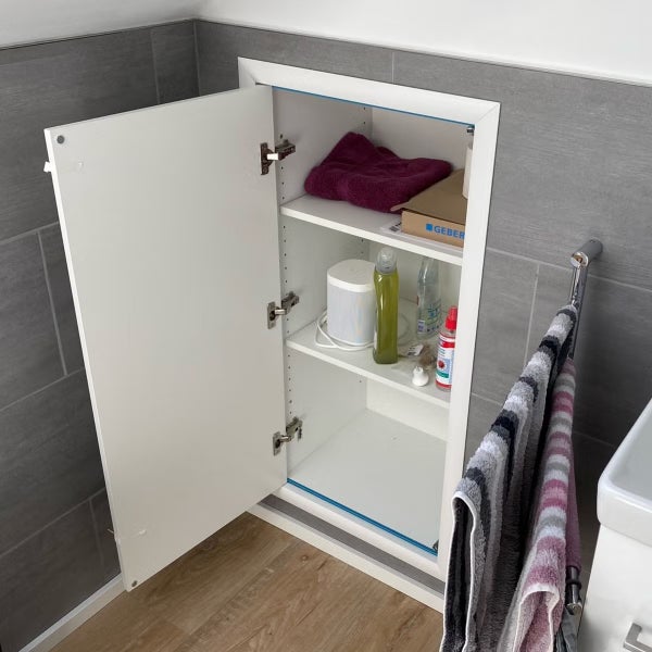 Ein weißer, in die Wand eingelassener Badezimmerschrank mit geöffneter Tür, montiert in einer gefliesten Wand mit Holzoptikboden.