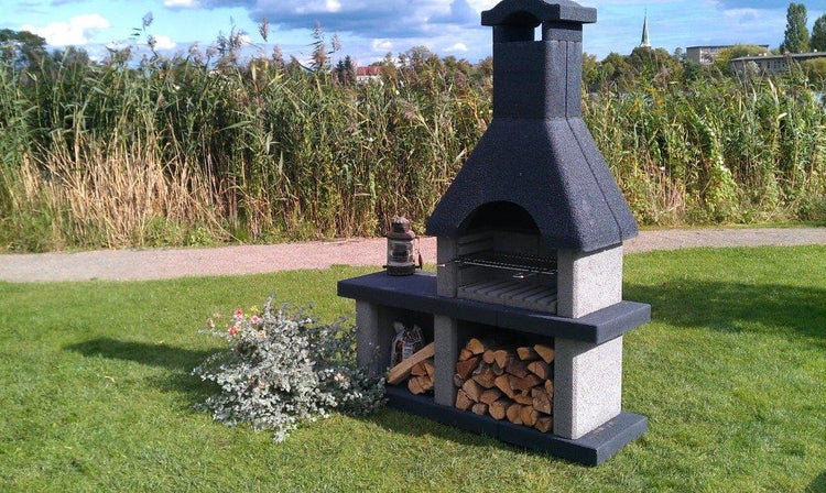Rustikaler Gartenkamin aus Stein mit Schornstein, Grillrost, seitlicher Ablage und Brennholzfach auf einer grünen Wiese.