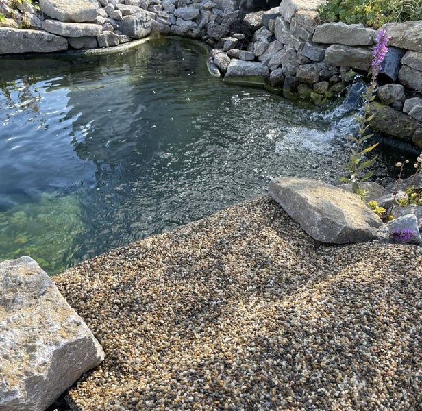 Gartenteich mit Natursteinumrandung, kleinem Wasserfall und einer mit Kies bedeckten Fläche im Vordergrund mit großen Ziersteinen.