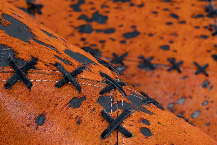 Detailaufnahme einer strukturierten Kuhfell-Oberfläche mit orangefarbenen und schwarzen Flecken sowie dekorativen schwarzen Kreuzstichen.