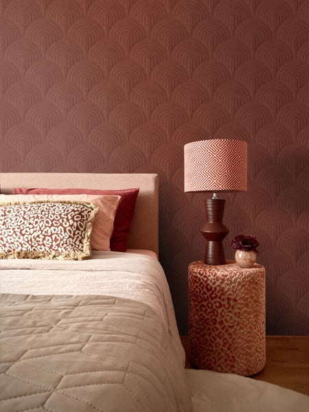 Schlafzimmer mit Tapete im Bogendesign, Bett mit dekorativen Kissen und Tischlampe auf einem gemusterten Pouf als Beistelltisch.