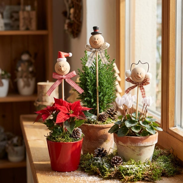 Drei winterliche Topfpflanzen mit dekorativen Holzfiguren auf Stäben, in Tontöpfen arrangiert als weihnachtliche Fensterdekoration.