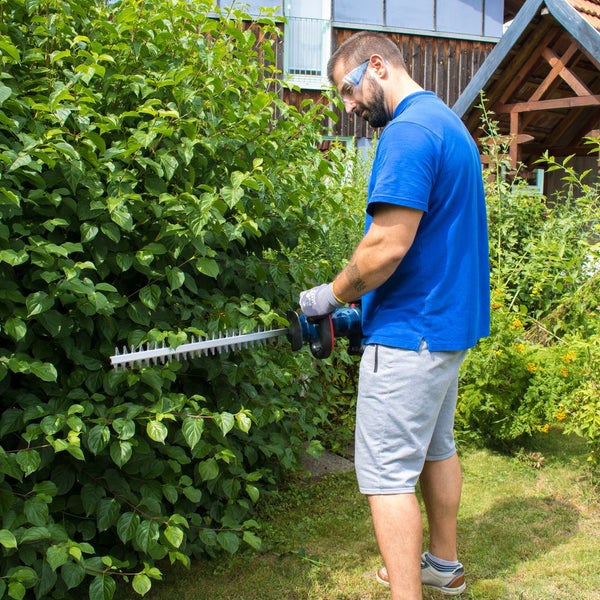 Ein Mann mit Schutzbrille und Handschuhen schneidet eine große grüne Hecke im Garten mit einer elektrischen Heckenschere.