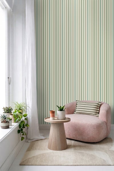 Vliestapete mit vertikalem Streifenmuster in einem hellen Wohnzimmer mit rosa Sessel, Holzbeistelltisch und verschiedenen Zimmerpflanzen.