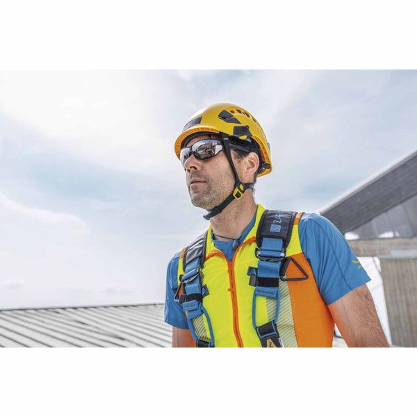 Mann mit gelbem Schutzhelm, Schutzbrille, Warnweste und blauem Sicherheitsgeschirr auf einer Baustelle.