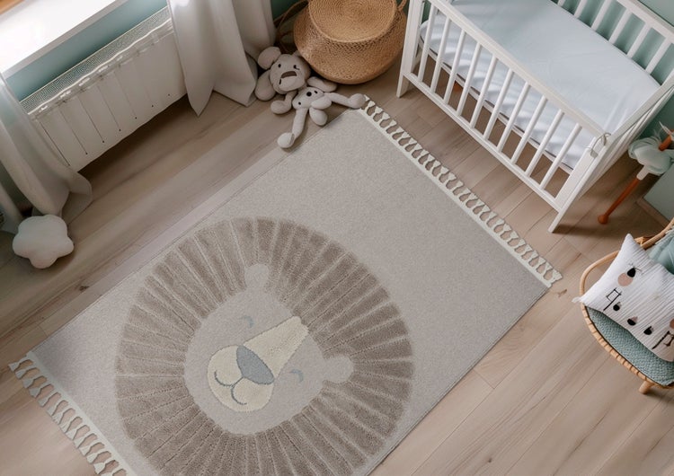 Ein Kinderteppich mit Löwenmotiv und Fransen im Kinderzimmer mit weißem Gitterbett, Spielzeug und hellen Holzdielen als Bodenbelag.