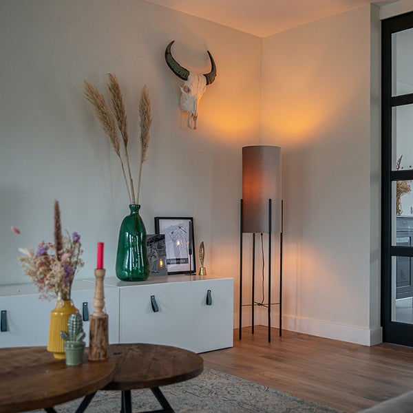 Modernes Wohnzimmer mit einer zylindrischen Stehlampe auf schwarzem Metallgestell, weißem Sideboard mit Glasvasen und einer Tierschädel-Wanddekoration.