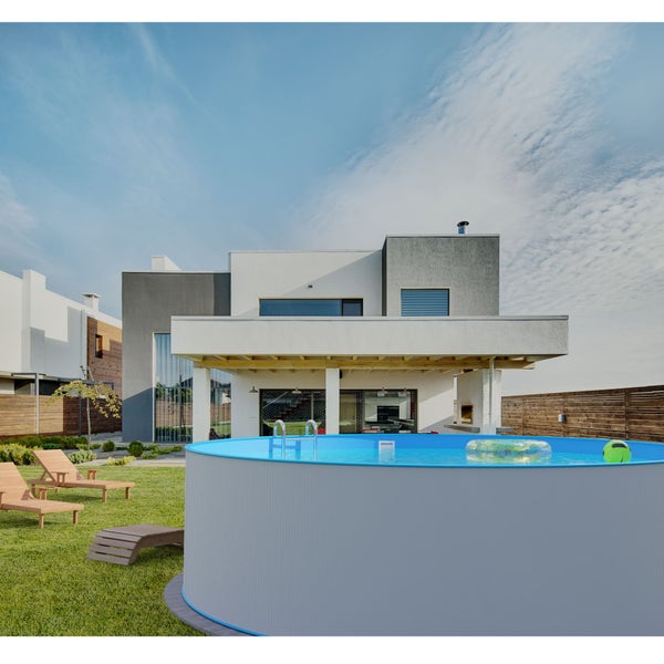 Runder Stahlwand-Aufstellpool mit Leiter in einem modernen Garten mit Rasen und hölzernen Sonnenliegen vor einem Wohnhaus.