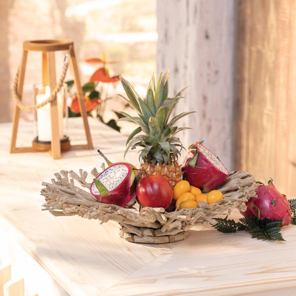 Dekorative Obstschale aus hellen Zweigen auf einem Holztisch, gefüllt mit tropischen Früchten wie Ananas und Drachenfrucht, neben einer Laterne.