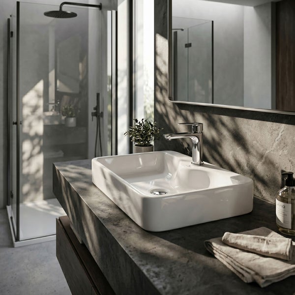 Modernes Badezimmer mit weißem Aufsatzwaschbecken aus Keramik auf einer dunklen Steinplatte, verchromter Armatur und Glasdusche im Hintergrund.