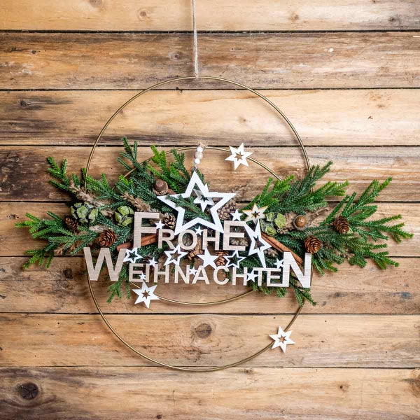 Weihnachtsdekoration mit Tannenzweigen, Zapfen, Sternen und dem Schriftzug Frohe Weihnachten an einem goldenen Ring.