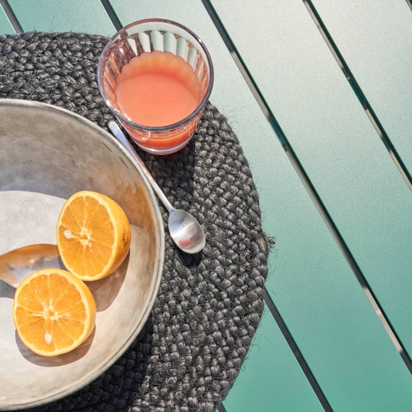 Gartentisch mit Metalllamellen, rundes geflochtenes Platzset, Keramikschale mit Zitronen und ein Glas Saft bei hellem Tageslicht.