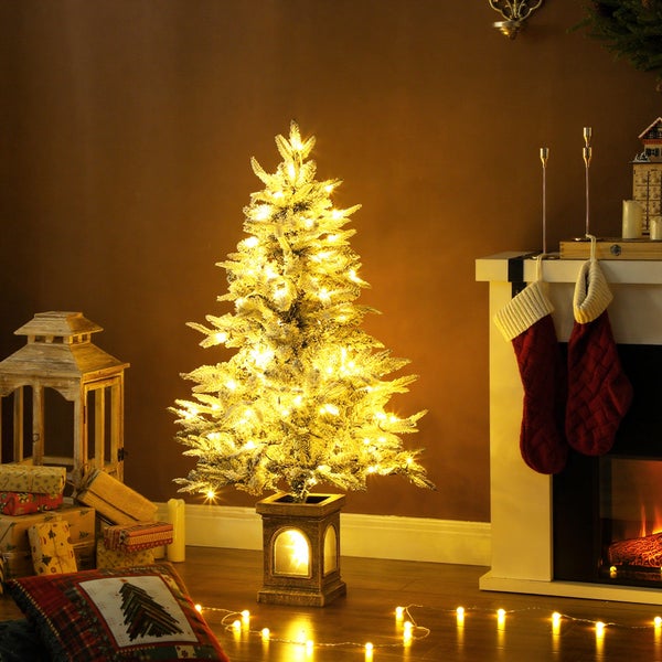 Kleiner, beschneiter Weihnachtsbaum mit warmweißer Beleuchtung in einem festlich dekorierten Wohnzimmer mit Kamin und Laterne.