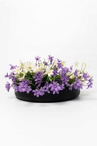 Künstliches Gesteck mit lila und weißen Blüten in einer flachen schwarzen Schale.