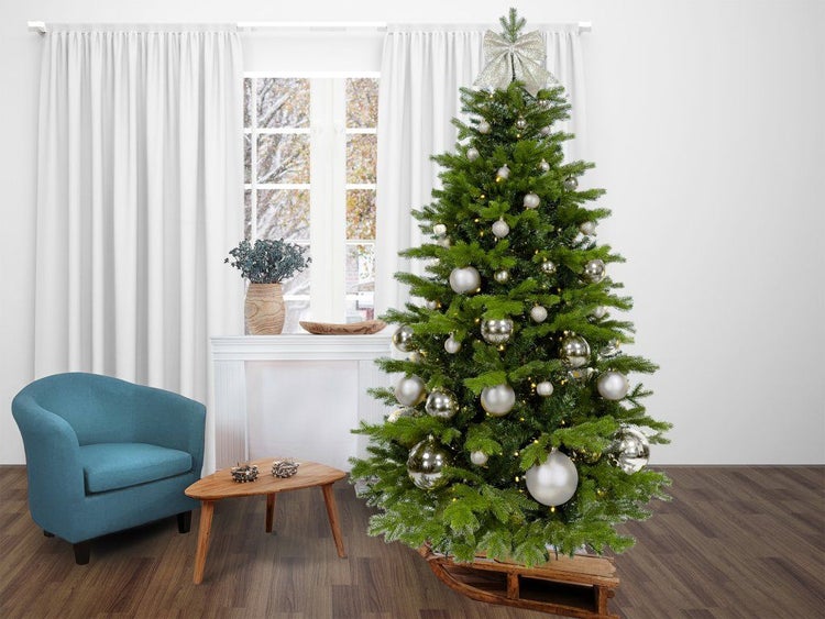 Geschmückter künstlicher Weihnachtsbaum mit Lichterkette in einem Wohnzimmer mit Sessel und Beistelltisch.
