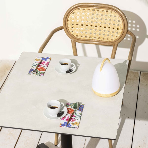 Quadratischer Bistrotisch, ein Rattanstuhl, Espressotassen auf Servietten und eine weiße Akkuleuchte mit Holzgriff auf einer Holzterrasse.