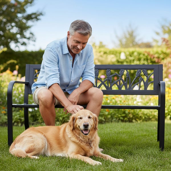 Ein Mann sitzt auf einer schwarzen Metall-Gartenbank mit Blumenmuster an der Lehne in einem hellen Garten, davor liegt ein Hund auf dem Rasen.
