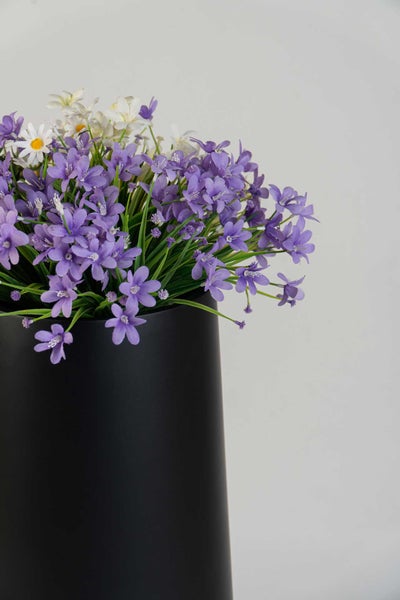 Künstliche lila und weiße Blumen in einer matten schwarzen Vase.