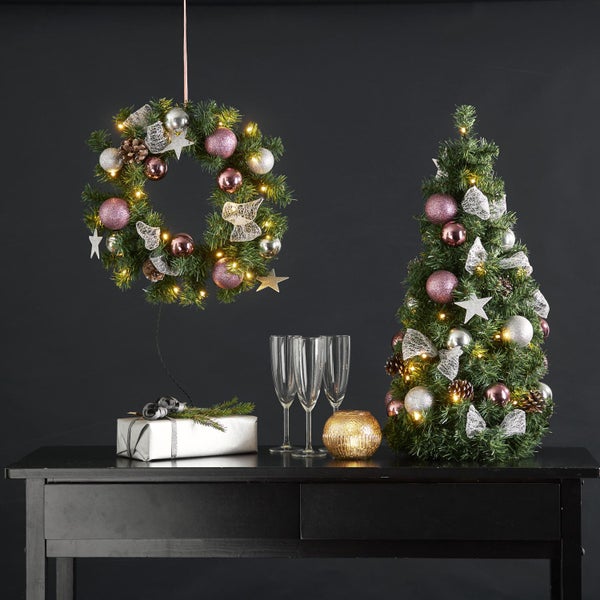 Dekoratives Arrangement mit Weihnachtskranz, Weihnachtsbaum, Geschenk und Gläsern auf einem Tisch.