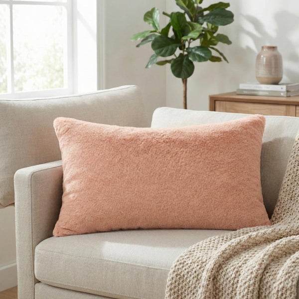 Rechteckiges, pfirsichfarbenes Plüsch-Dekokissen auf einem hellen Sofa in einem hellen Wohnzimmer mit Pflanze und Strickdecke.