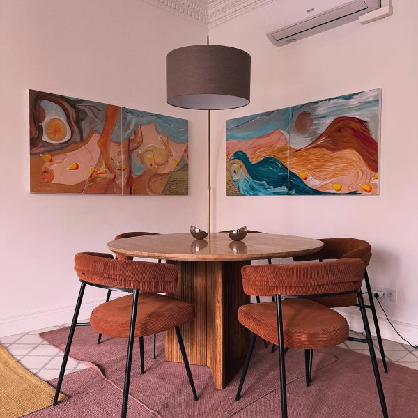 Esszimmer mit rundem Holztisch, vier Stühlen mit Bezug in Cordoptik, einer Stehleuchte und zwei großen abstrakten Wandgemälden in einem hellen Raum.