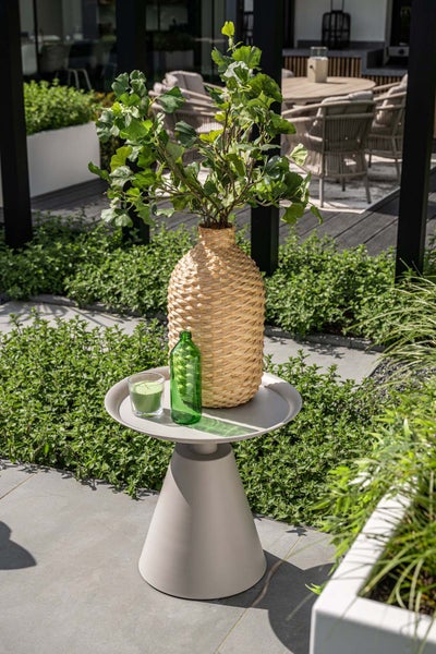 Heller, organisierter Gartenbereich mit modernem Beistelltisch mit kegelförmigem Fuß, geflochtener Vase mit Zweigen, Glasflasche und Kerze.