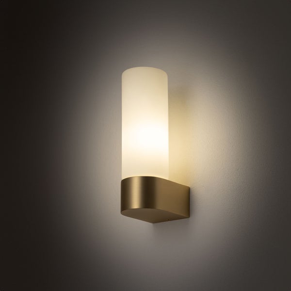 Moderne Wandleuchte mit zylindrischem Schirm aus satiniertem Glas und goldfarbenem Metallsockel, eingeschaltet mit warmem Licht.