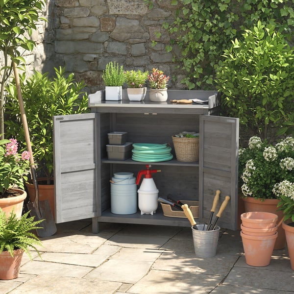 Grauer Garten-Pflanztisch aus Holz mit zwei offenen Türen und Metallarbeitsplatte, gefüllt mit Gartenzubehör in einem organisierten Garten.