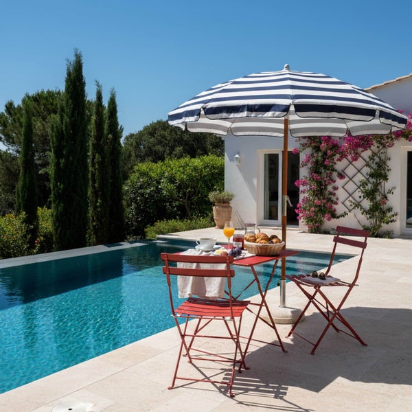 Mediterrane Poolterrasse mit einer roten Garten-Sitzgruppe aus Metall und einem gestreiften Sonnenschirm neben einem Swimmingpool.